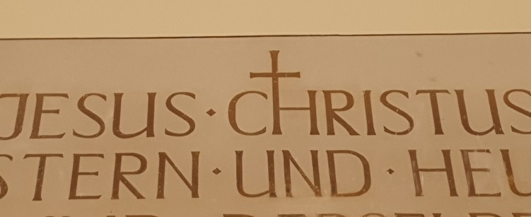 A picture of an insignia in a church spelling the words “Jesus Christ” and other text below it. The H is extended upwards in order to use one one of the vertical lines for the drawing of a cross. I am making fun of that tact as the letter “T” already looks like a cross, so why would they go the extra mile and make the H look like one? This doesn't make any sense.