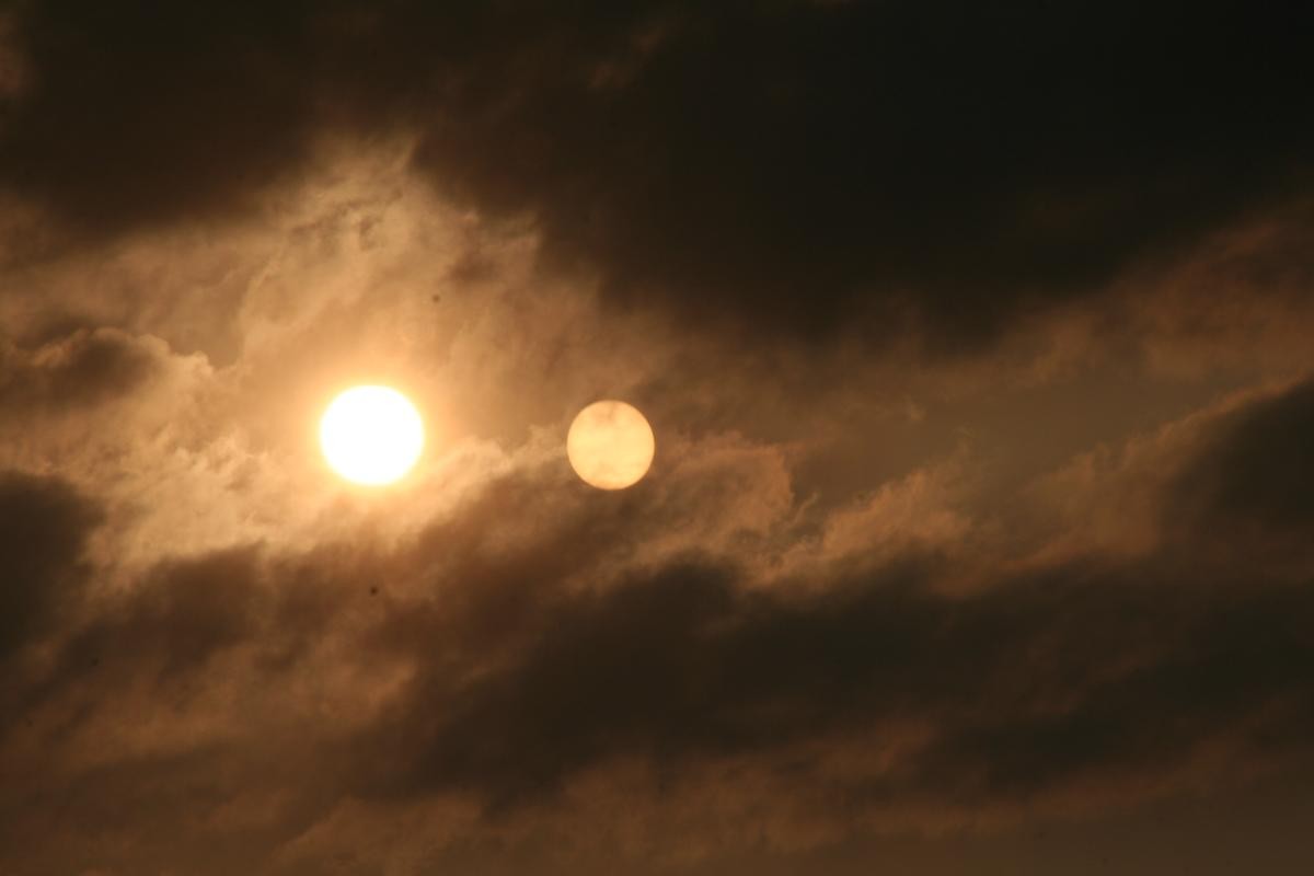 Foto im Querformat. Im roten Abendlicht mit dicken Wolken blickt die Kamera direkt in die Sonne. Ein Stück rechts daneben scheint eine zweite, gleich große, aber blassere Sonne zu stehen.