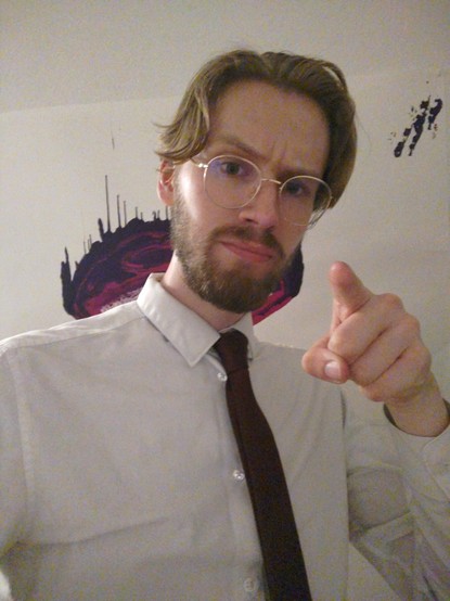A picture of Erik Uden pointing into the camera. He's wearing a tie and making a serious face. Behind him is a picture he drew in the KRH Wunstorf