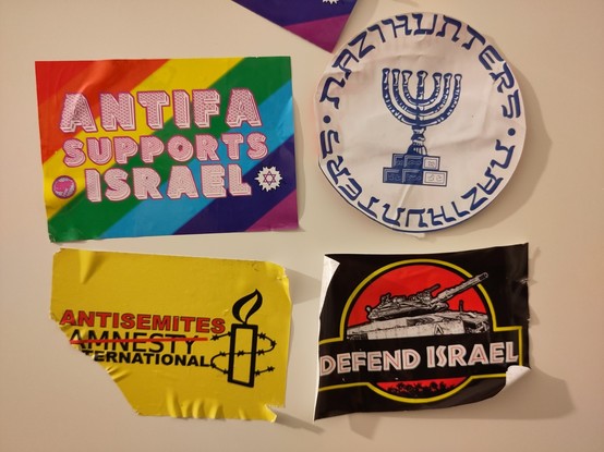 Four stickers on a wall saying:

1 “Antifa supports Israel” with a tilted rainbow flag in the whole background, an entirely red Antifa flag logo in the bottom left, and a star of David with a white background in the bottom right

2 the Mossad logo (Israel's secret service) but instead of Hebrew text it says “Nazihunter” twice

3 „Antisemites International” with the Logo of Amnesty International (a candle with barbed wire surrounding it) but the word Amnesty crossed out and replaced with “Antise…
