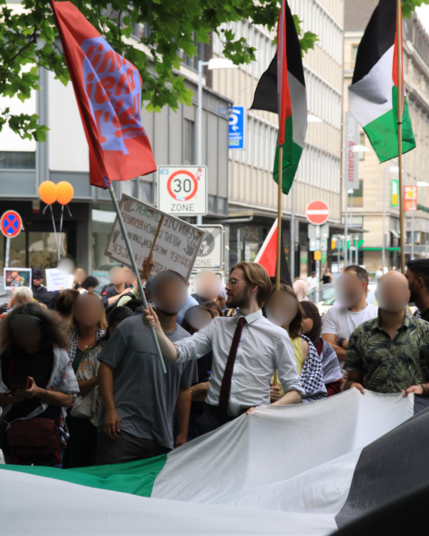 Ein Bild von Erik Uden in einer Menschenmenge draußen auf deutschen Straßen. Alle Gesichter sind zensiert außer das von Erik. Man sieht wie er eine rot-blaue Linksjugend ['solid] Flagge trägt und zwei weitere Personen Palästina Flaggen. Auf dem Bild sind ungefähr 20 Menschen zu sehen. Oben rechts im Hintergrund sieht man zwei Orange Ballons welche von der Gegendemo einer Pro-Israel Organisation namens „Iron Dome Hannover” stammen.

Es tragen viele Menschen eine Kufiya und oder Hijab. 

Das Bild…