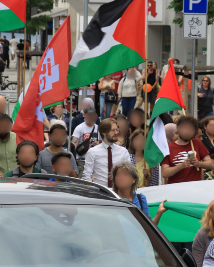 Ein Bild von Erik Uden in einer Menschenmenge in der jedes Gesicht zensiert ist außer das eigene. Eine Person links neben ihm hält eine rot-blaue Linksjugend ['solid] Flagge, zwei Personen rechts neben ihm tragen eine Palästina Flagge. Im Hintergrund sind weitere Menschen mit orangen Ballons zu erkennen, welche von der „Iron Dome Hannover” Gegendemo kommen. Auf dem Bild sind ungefähr 30 Leute zu erkennen.

Es tragen viele Menschen eine Kufiya und oder Hijab. 

Das Bild wurde aufgenommen am Sams…