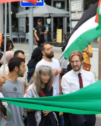 Ein Bild von Erik Uden in einer kleinen Menschenmenge von ungefähr 10 Personen. Auf dem Bild hält er eine nicht im Bild erkennbare sehr große Palästina-Flagge und spricht mit einer Person links neben ihm.

Es tragen viele Menschen eine Kufiya und oder Hijab.

Das Bild wurde aufgenommen am Samstag den 28.06.2025 nach 17:00 Uhr in Hannover.