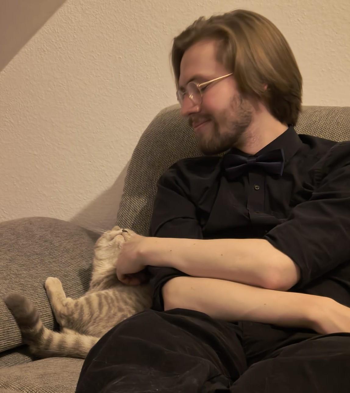 An image of Erik Uden petting his cat Lenin whilst both are sitting on a couch. Lenin is smiling with enthusiasm and moving his head towards Erik's hand to signal that he likes being pet.