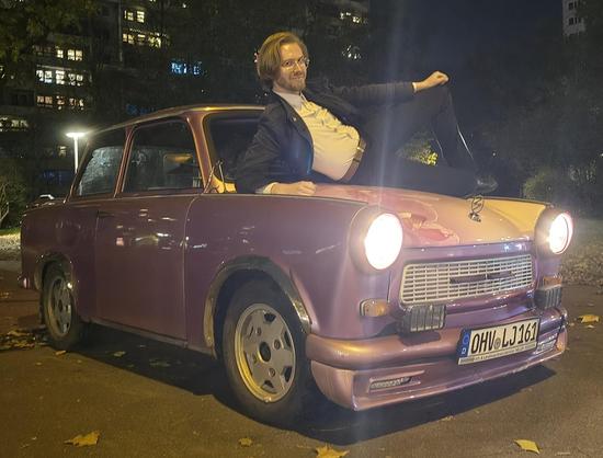 Ein Bild von Erik Uden wie er auf der Motorhaube eines pink lackierten Trabants mit dem Nummernschild „LJ 161” (Linksjugend Antifaschistische Aktion) posiert, in Berlin Friedrichshain vor dem Franz-Mehring-Platz 1, dem Gebäude der Zeitung Neues Deutschland.