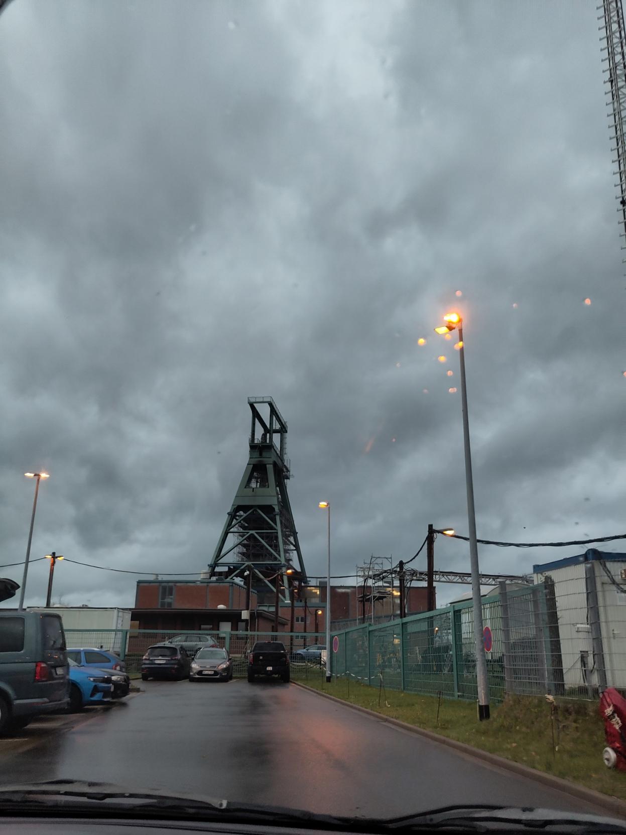 An image of the Konrad nuclear waste storage facility from the outside at 8 AM during rain. The big green tower is what pulls the elevator up and down, and it's stationed within buildings with red bricks. In front of all of this we can see a parking lot. This image was taken from the insides of a car.
