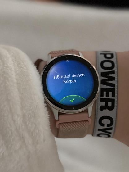 Der Schriftzug „Höre auf deinen Körper“ wird auf einer Smartwatch angezeigt. Die Smartwatch hängt an einem Handgelenk links von einem 39c3-Bändchen.
