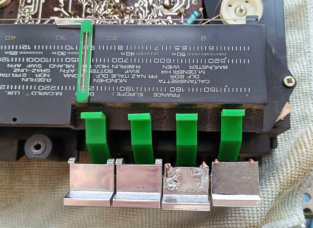 Teil eines Radios, ma sieht 4 Tasten in chrom, die in Reparatur sind. 
Die beiden linken Tasten sind Ok, die beiden rechten Tasten sind : Mitte, noch stark beschädigt, rechts ist ein Stück Aluminium aufgeklebt.
Das neue Aluminium zeichnet sich durch eine feine, bürstete Struktur aus.