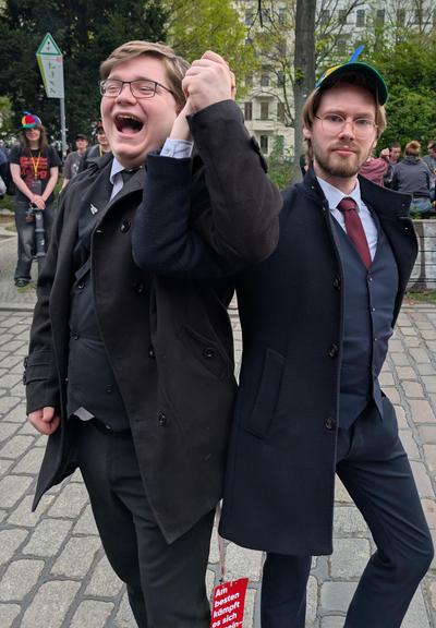 A picture showing Julian Schlingmann and Erik Uden holding hands whilst standing and posing back to back. Both are wearing a professional multi layered suit, yet only Erik Uden is wearing a children's propeller hat. In the background on the left you can see another person wearing such a propeller hat. They are outside standing on the street. This picture was taken at the bUm.