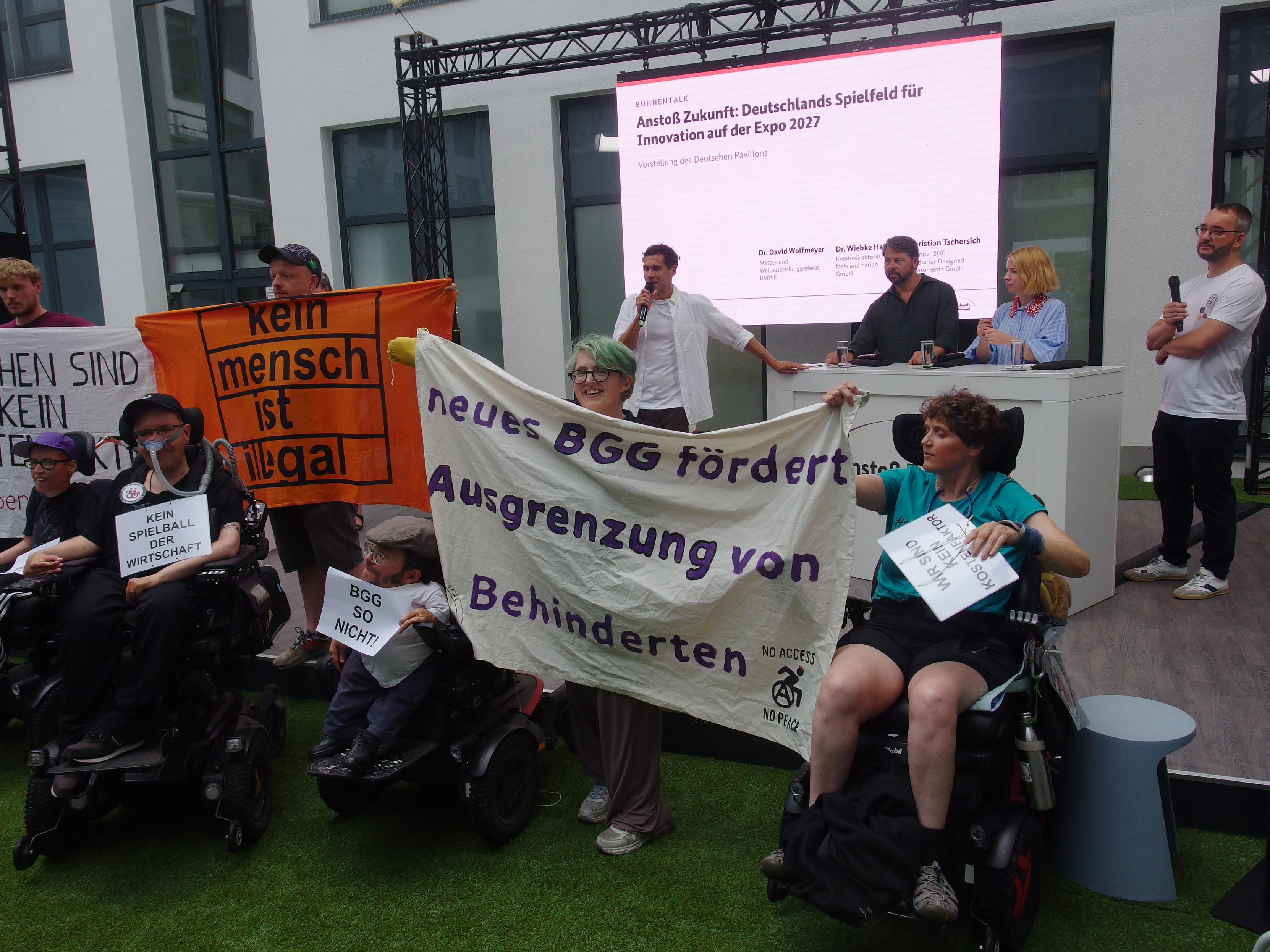 Protest vor der Bühne. im hintergrund das programm, mit Powerpoint und Speakers. Davor die Aktivistis mit Bannern: neues BGG fördert Ausgrenzung vin Behinderten, kein mensch ist illegal