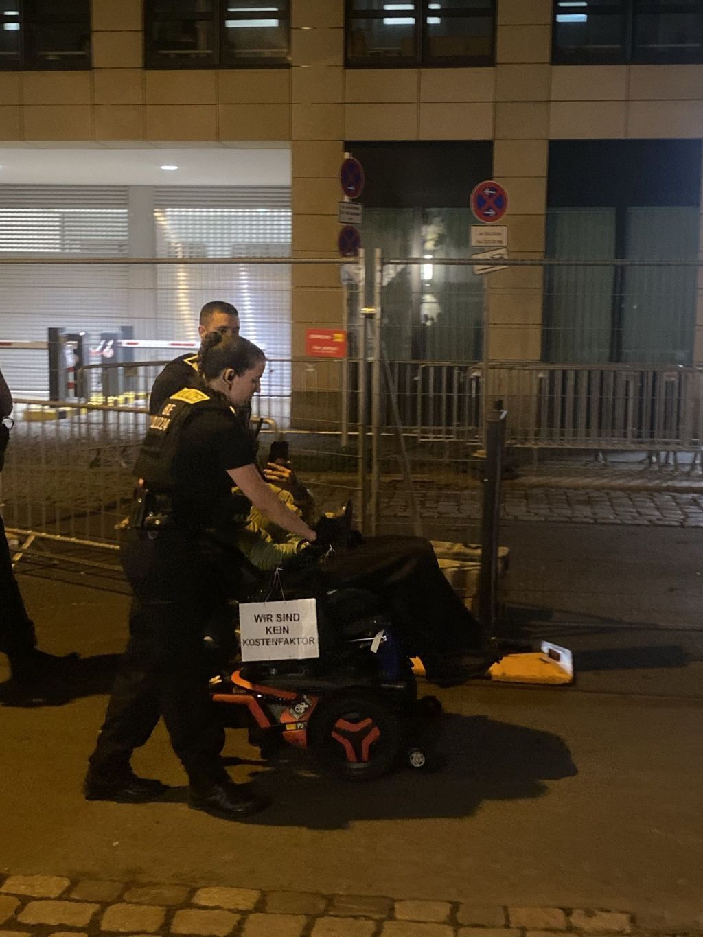 eine Person im E-Rollstuhl wird von der Polizei begleitet, diese steuert den rollstuhl. die Aktivistin hat ein Schild "wir sind kein kostenfaktor"