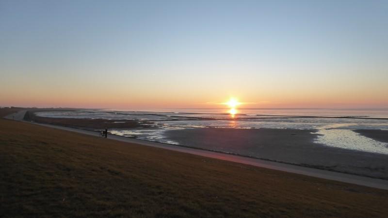 Sonnenuntergang über der Nordsee. Im Vordergrund der Deich, dann diagonal von linls oben nach rechts unten Watt mit Prielen. Die untergehende Sonne direkt auf dem Horizont in der Bildmitte beleuchtet die Atmosphäre zu einem orangenen Streifen über die gesamte Breite. Darüber in unterschiedlichen Blautönen der wolkenlose Himmel