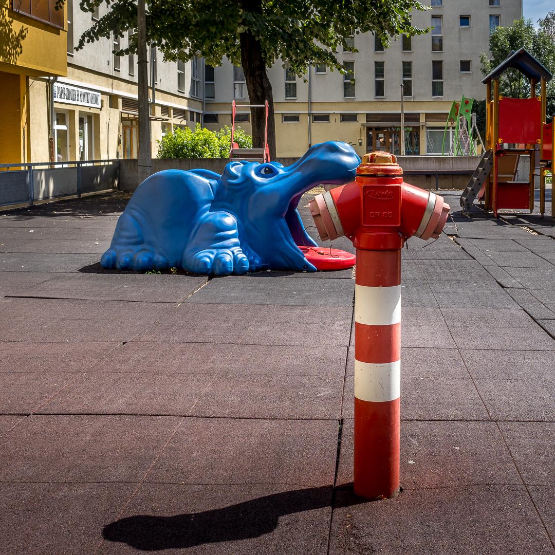Ein Spielplatz inmitten eines Wohngebietes. Im Vordergrund ein leuchtend roter Hydrant. Dahinter links eine Nilpferdrutsche in blau mit weit aufgesperrtem Maul, perspektivisch frisst es den Hydranten.