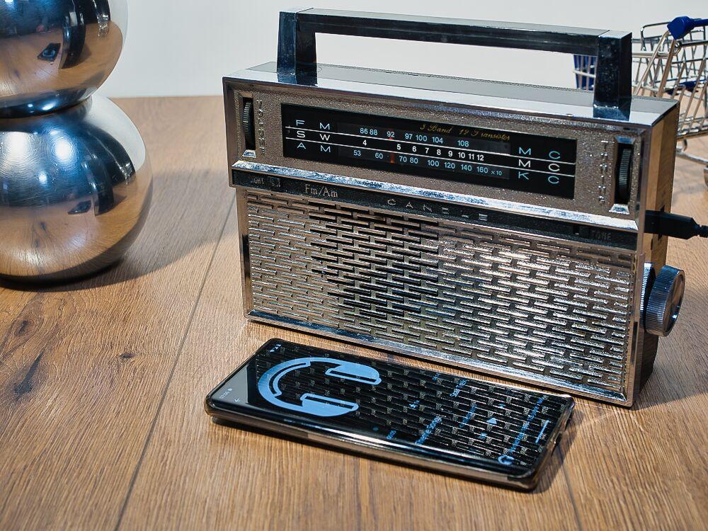 Ein chromfarbiges Kofferradio vor dem ein Smartphone liegt.
Auf dem Bildschirm des Smartphones ist ein Kopfhörer zu sehen.
Links sind 2 chromfarbene Bälle übereinander.