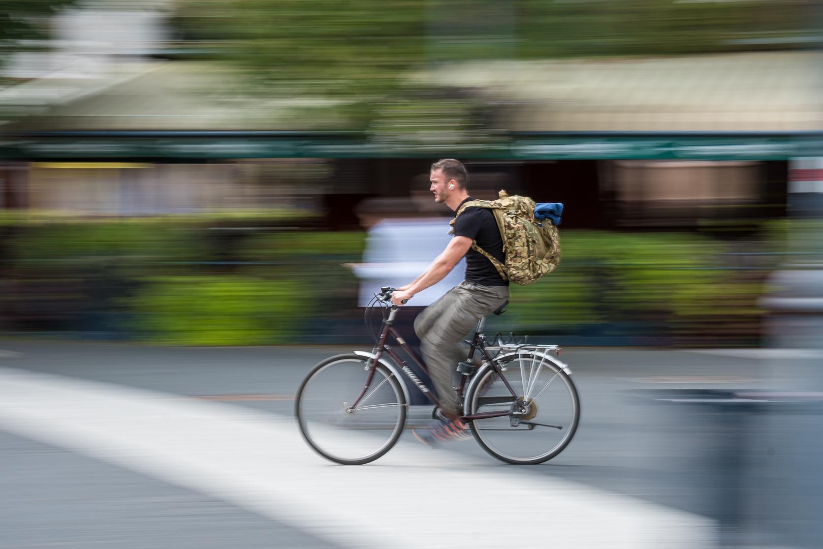 Ein Radfahrer mit großem Tarnrucksack rast durch die Stadt. Durch das Mitziehen der Kamera verschwimmt der Hintergrund zu horizontalen Streifen, während Fahrer und Fahrrad weitgehend scharf bleiben.