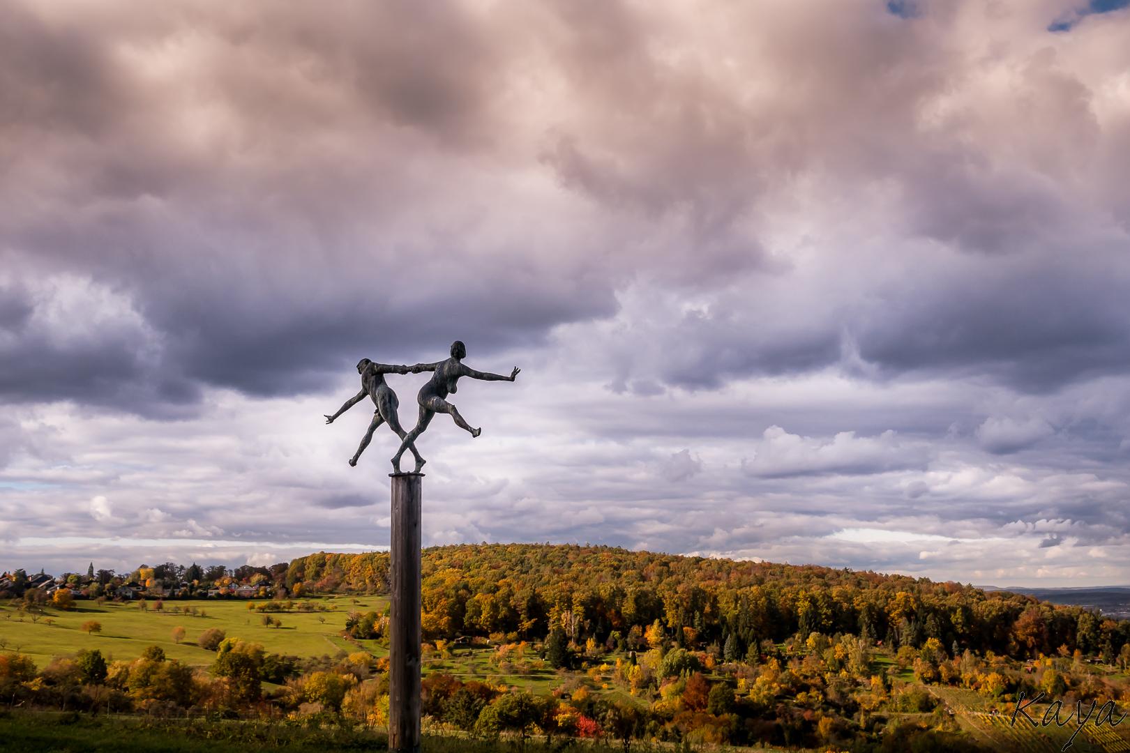 Zwei Figuren balancieren gemeinsam auf einer hohen Säule über einer herbstlichen Landschaft, während schwere Wolken über den Hügeln ziehen.