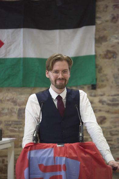 Ein professionelles Bild von Erik Uden in Hemd, Weste und Krawatte wie er vor einem Rednerpult steht und in die Kamera lächelt. Hinter ihm eine Flagge des Staates Palästina.