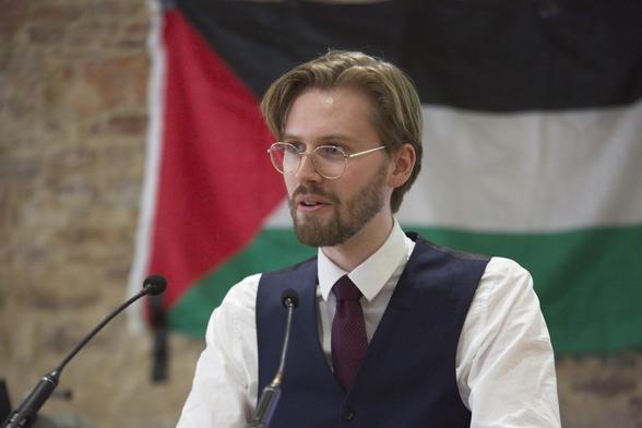 Ein professionelles Bild von Erik Uden in Hemd, Weste und Krawatte wie er vor einem Rednerpult steht und zur Seite schaut. Hinter ihm eine Flagge des Staates Palästina.
