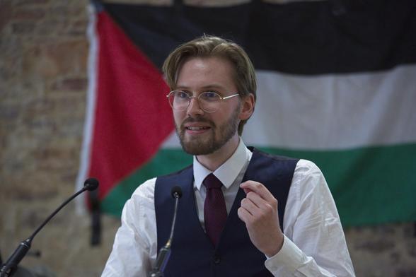 Ein professionelles Bild von Erik Uden in Hemd, Weste und Krawatte wie er vor einem Rednerpult steht und seine Hand hebt wie er etwas erklärt. Hinter ihm eine Flagge des Staates Palästina.